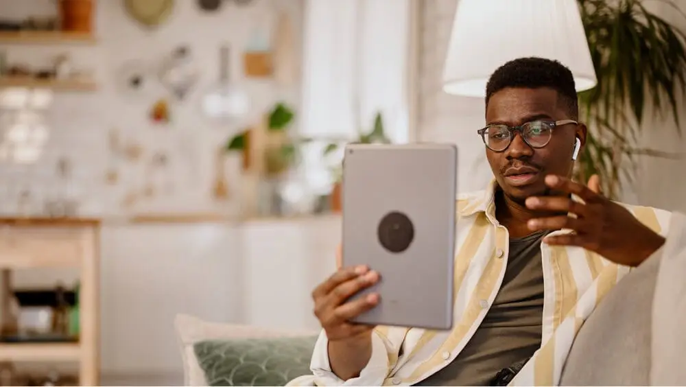 A man sitting comfortably on a couch at home, holding an iPad and participating in a video meeting for online therapy.